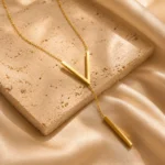 Gold-plated stainless steel Vertex V Lariat Necklace with geometric bar pendant on beige stone background