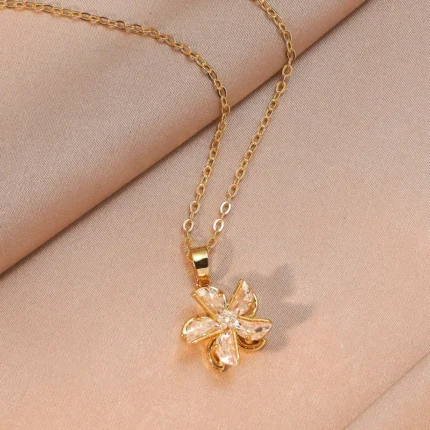 Gold-plated stainless steel necklace featuring a swirling five-petal flower pendant with clear faceted stones on a pink satin background.