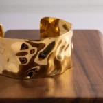 Wide gold-plated stainless steel cuff bracelet with a hammered, undulating texture resting on a wooden surface.