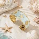 Gold-plated cuff bracelet with light blue enamel, starfish, and shell motifs on sand
