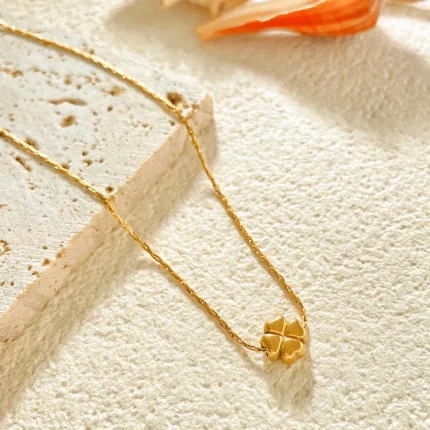 Gold-plated stainless steel necklace with a small four-leaf clover pendant on a textured stone surface.