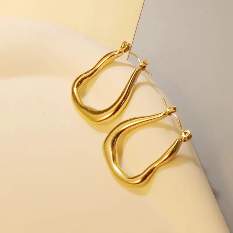 Gold-plated stainless steel hoop earrings with a wavy, undulating frame design displayed on a textured stone surface.
