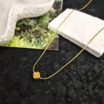 Gold-plated stainless steel necklace with a small four-leaf clover pendant on a textured stone surface.