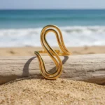Gold and silver stainless steel S-curve statement rings worn on a hand and displayed on a beach log.