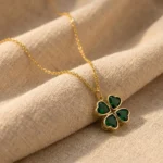 Gold-plated stainless steel four-leaf clover pendant necklace with emerald green heart-shaped stones on lace background