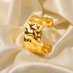 Wide gold-plated stainless steel cuff bracelet with a hammered, undulating texture resting on a wooden surface.