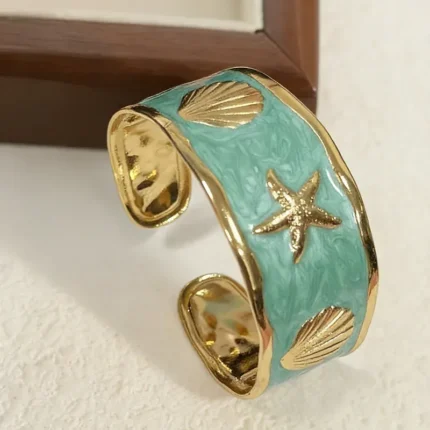 Gold-plated cuff bracelet with light blue enamel, starfish, and shell motifs on sand