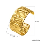 Wide gold-plated stainless steel cuff bracelet with a hammered, undulating texture resting on a wooden surface.
