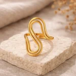 Gold and silver stainless steel S-curve statement rings worn on a hand and displayed on a beach log.