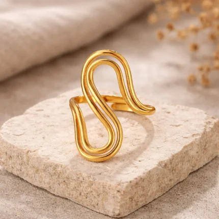 Gold and silver stainless steel S-curve statement rings worn on a hand and displayed on a beach log.