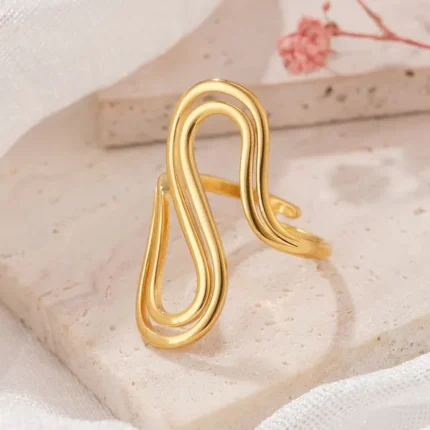 Gold and silver stainless steel S-curve statement rings worn on a hand and displayed on a beach log.